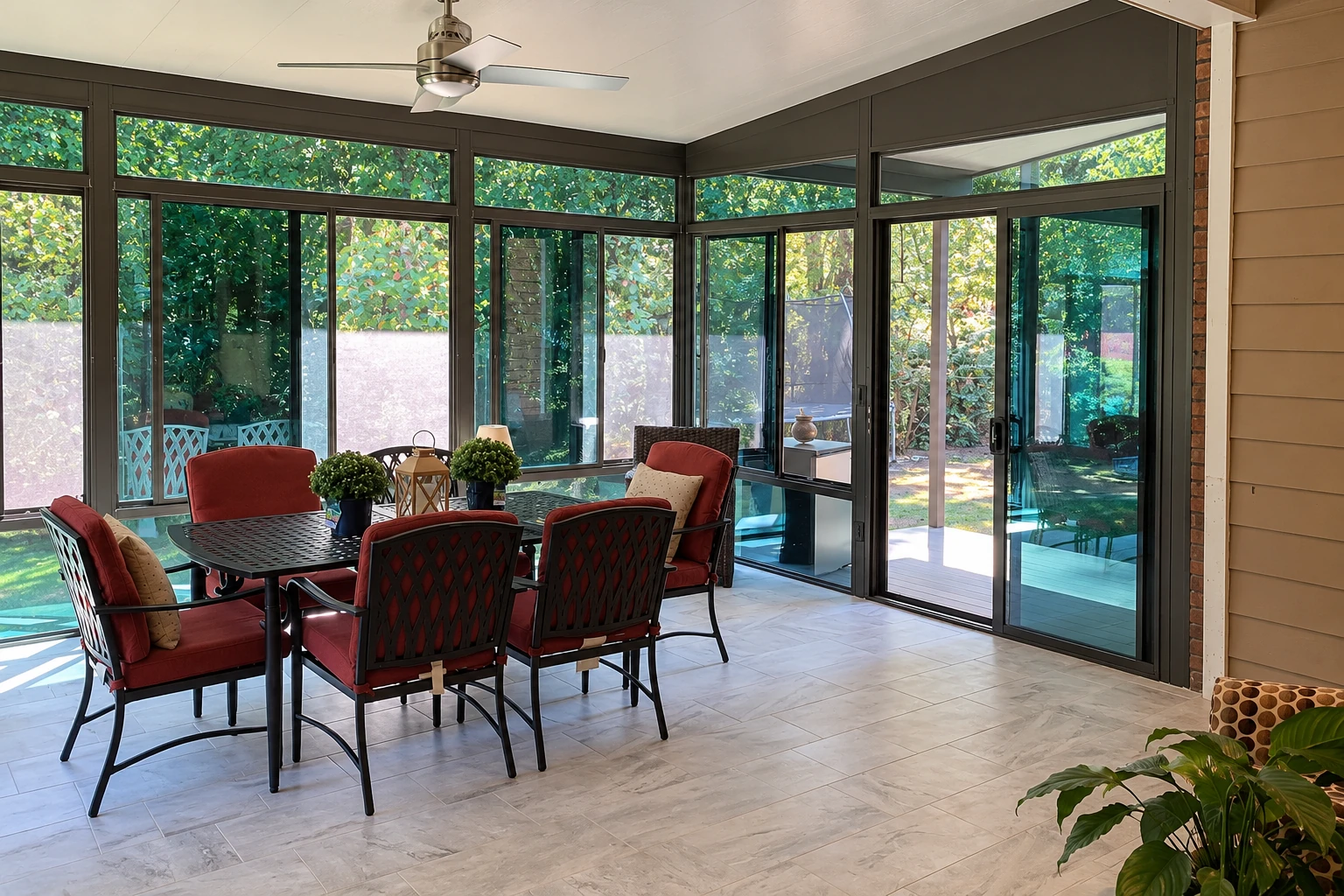 A glass-enclosed patio room has dark frames, tile flooring, a ceiling fan, and a dining table with red-cushioned chairs.