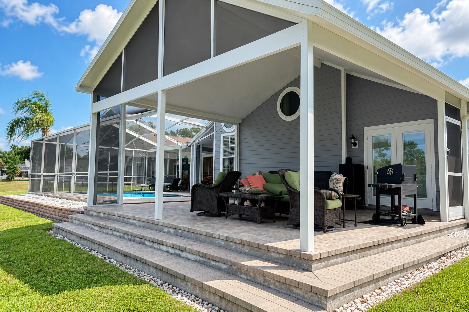 A white patio cover shelters outdoor seating beside a gray house, with a screened enclosure and pool visible beyond.