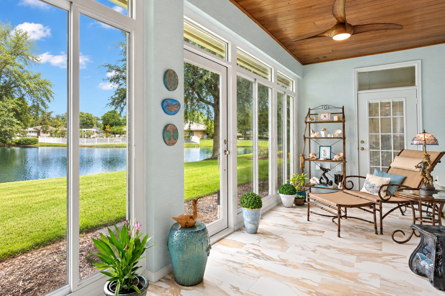 Bright sunroom with white-framed windows overlooking a lake, furnished with a wooden lounge set and potted plants.