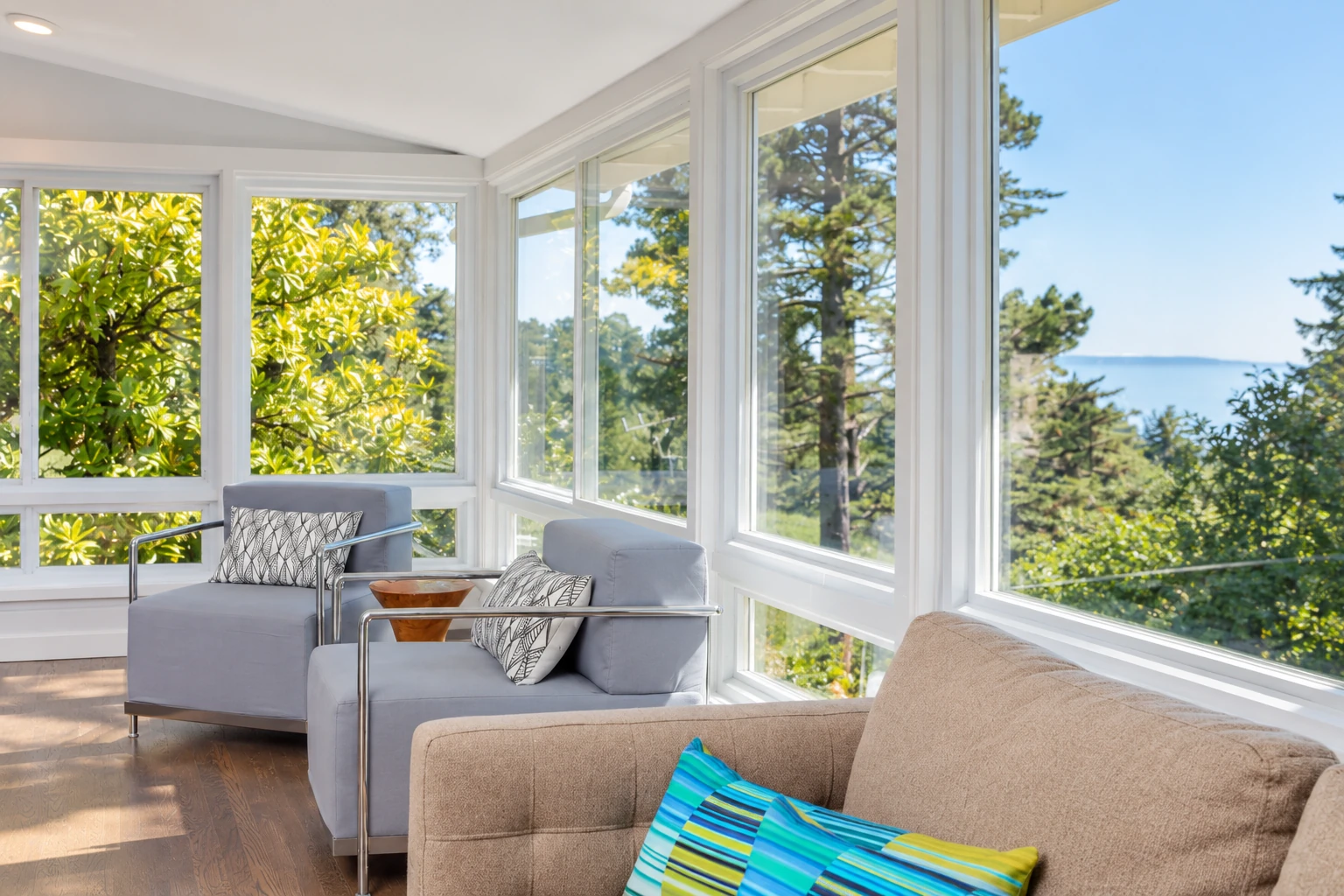 Sunroom with expansive windows overlooking trees and water, furnished with gray and beige sofas.