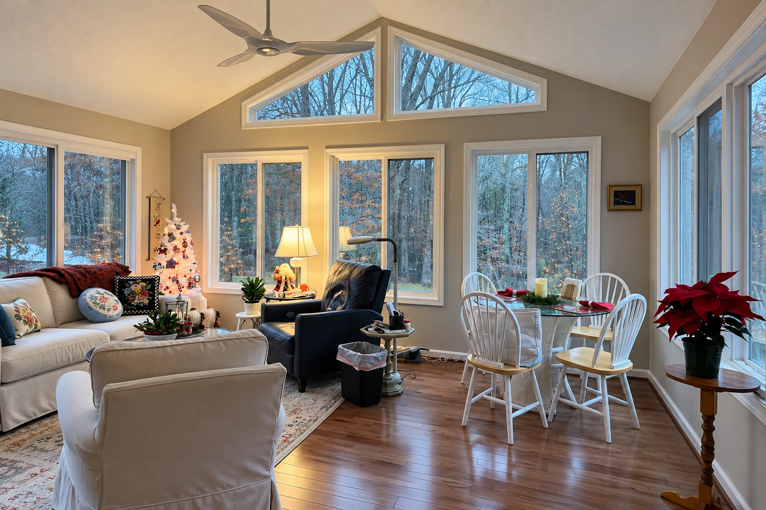 A furnished sunroom with large windows, a dining set, ceiling fan, and a small decorated Christmas tree.