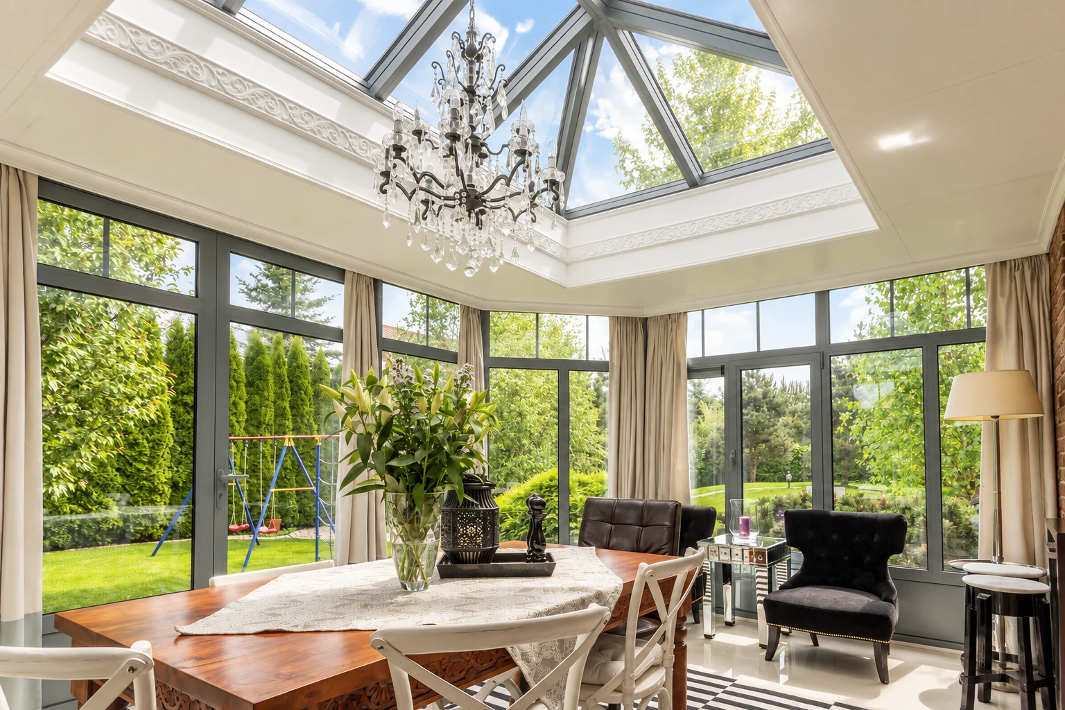 Bright sunroom with a glass roof, tall windows, dining table and chairs, and a chandelier.